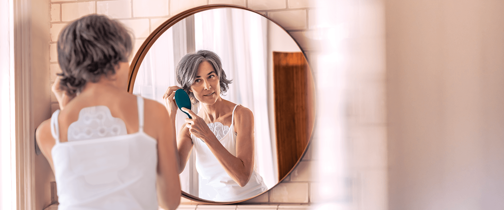 Woman wearing Optune Lua arrays on her back and chest brushes her hair in the mirror.